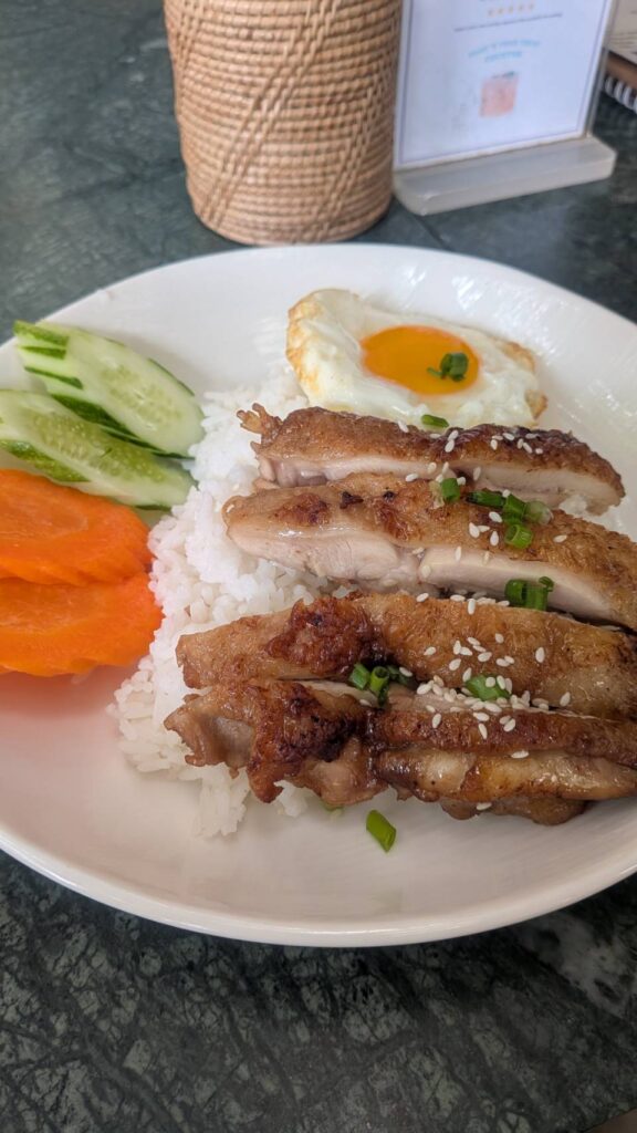 白いお皿に盛られたバイサチュルー。ご飯の上に焼いた豚肉と目玉焼き、きゅうりとにんじんの付け合わせ